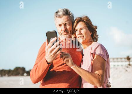 Smiling mature couple using mobile phone at beach Stock Photo