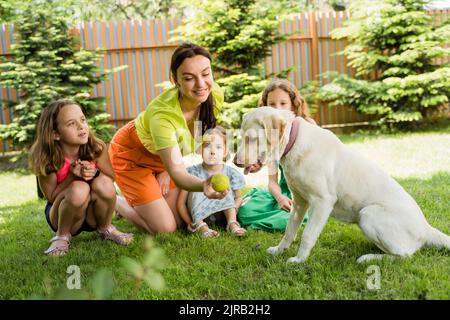 Smiling woman showing ball to dog by daughters at back yard Stock Photo