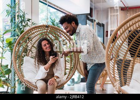 Businessman sharing mobile phone with businesswoman sitting in hanging chair Stock Photo