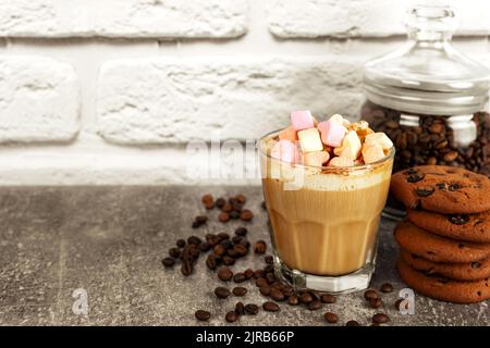 Caramel latte coffee in a glass with marshmallow top and chocolate cookie. Coffee beans on gray and white brick wall surface with copy space. Stock Photo