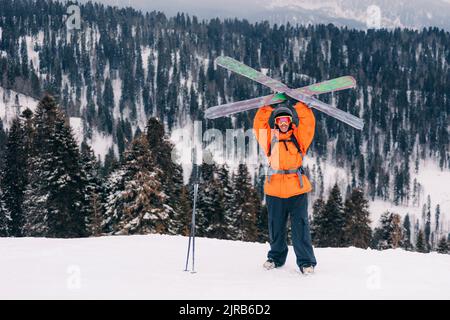 Smiling skier holding skis standing with arms raised in front of mountains Stock Photo