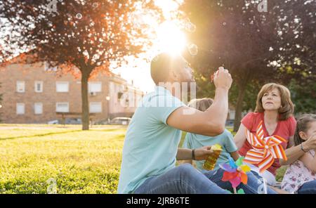 Man blowing bubbles sitting by family at park Stock Photo