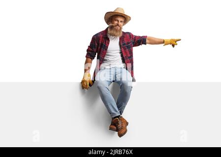 Full length portrait of a young farmer with gloves and a straw hat sitting on a panel and pointing isolated on white background Stock Photo