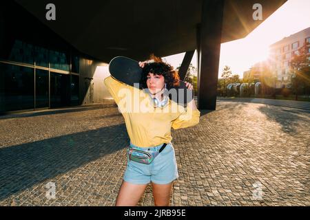 Young woman with skateboard standing under bridge at sunset Stock Photo