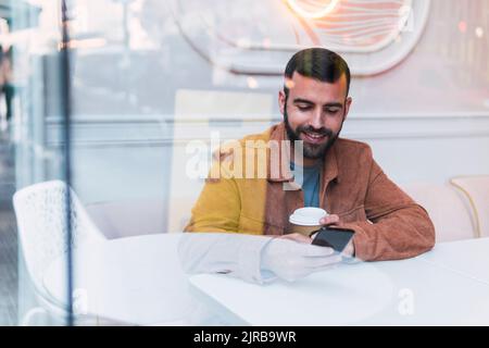 Smiling young man using smart phone seen through glass at cafe Stock Photo