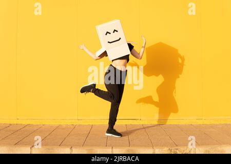 Playful woman wearing box with smiley face gesturing in front of yellow wall on footpath Stock Photo