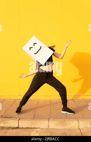 Playful woman wearing box with smiley face doing robot dance in front of of yellow wall Stock Photo