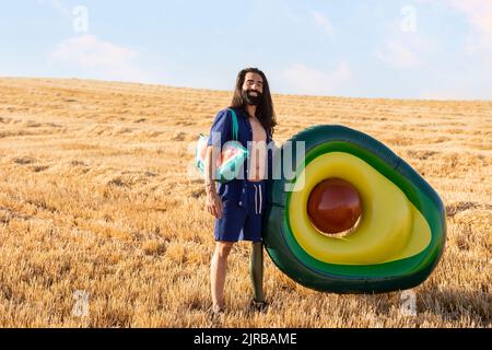 Smiling bearded man with prosthetic leg carrying bag by avocado shaped inflatable ring at field Stock Photo