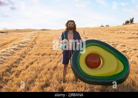 Bearded man with prosthetic leg wearing sunglasses standing by avocado shaped inflatable ring at field Stock Photo