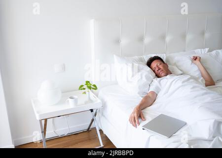 Mature man sleeping by laptop in bed Stock Photo