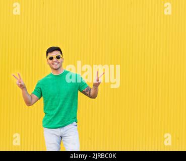 Happy young man gesturing peace sign in front of yellow wall Stock Photo