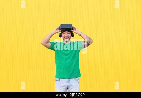 Happy young man standing with virtual reality simulator against yellow background Stock Photo