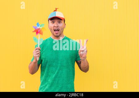 Young man with rainbow pinwheel toy showing peace sign in front of wall Stock Photo