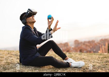 Young businessman with virtual reality simulator holding globe sitting on hill Stock Photo