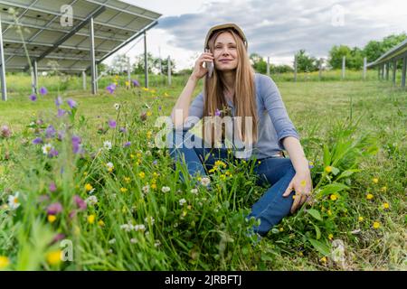 Smiling engineer talking on smart phone sitting in front of solar panels at field Stock Photo