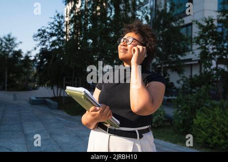 Smiling young businesswoman with eyes closed holding laptop talking on mobile phone Stock Photo