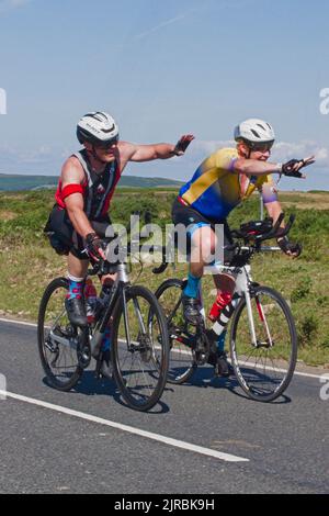 7thAug22 Gower Swansea Wales UK Triathlon Ironman cycling event Pair of cyclists side by side both waving to supporting spectators off camera. Stock Photo