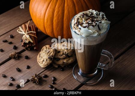 Glass of coffee with whipped cream, chocolate sprinkles. Autumn composition with cinnamon sticks, star anise, pumpkin, chocolate chip cookies, beans. Stock Photo