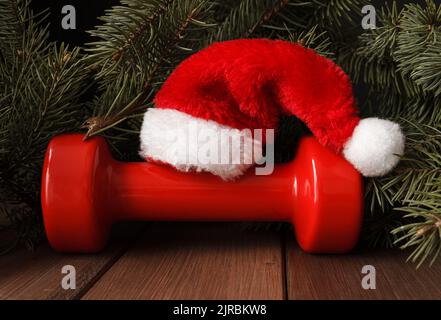 Red dumbbell with Santa Claus hat. Exercise equipment as Christmas gift idea. Gym fitness holiday season concept composition with tree branches. Stock Photo