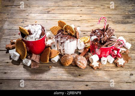 Christmas styled overloaded hot chocolate mugs with various toppings – chocolate slices, spices, sweets, cookies, candy and gingerbreads, wooden cozy Stock Photo