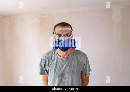 Selective focus, medium shot of sweaty caucasian man in safety glasses and dust mask, looking directly into the lens seriously. Stock Photo