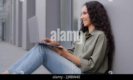 Smiling happy lady with long hair sitting on floor typing browsing laptop casual chatting woman user worker freelancer businesswoman leaning wall Stock Photo