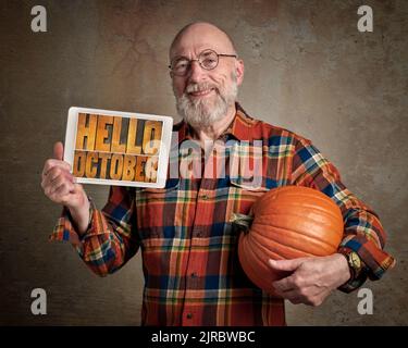 Hello October - smiling and cheerful senior with a big pumpkin and a digital tablet with  text in letterpress wood type, greeting card concept Stock Photo