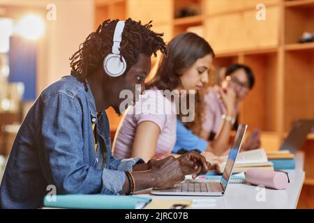 Diverse group of students in row using laptops and studying in college library, focus on black young man wearing headphones in foreground Stock Photo