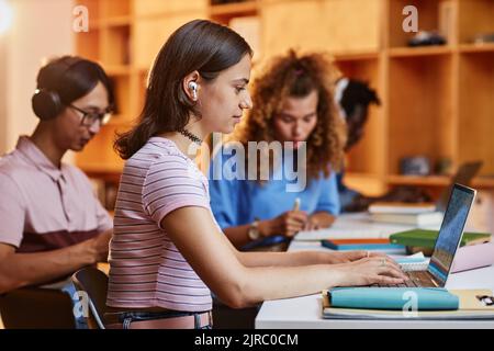 Diverse group of students in row using laptops and studying in college library, focus on smiling young woman wearing headphones in foreground Stock Photo