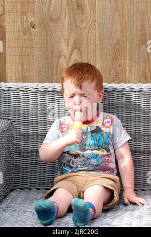 A young child is shown licking a sour flavoured ice lolly. The boy is sitting outside and is grimacing ts the bitter taste of the flavoured ice. Stock Photo