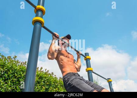 Muscular man in a cap doing pull-ups on the horizontal bar in the park outdoors Stock Photo