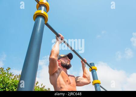 Muscular man in a cap doing pull-ups on the horizontal bar in the park outdoors. Stock Photo