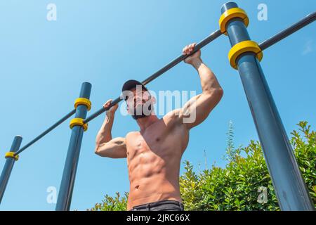 Muscular man in a cap doing pull-ups on the horizontal bar from behind his head in the park outdoors Stock Photo