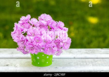 A bouquet of delicate pink carnations in a green decorative bucket on a white table against a blurred background of nature. Place for an inscription Stock Photo