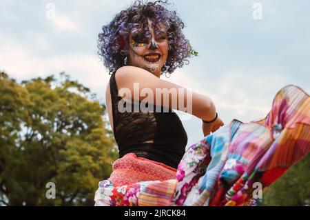 Latin young woman with La Calavera Catrina makeup, dressed in a long skirt outdoors dancing traditional dances. Stock Photo