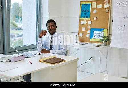 Portrait of young black teacher smiling at camera while sitting at desk in empty classroom, copy space Stock Photo