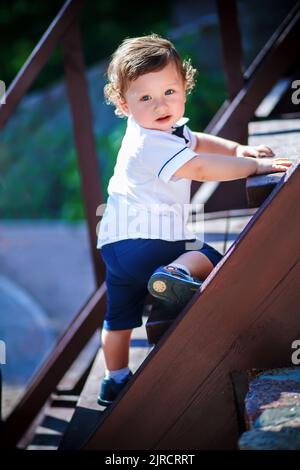 Little handsome boy climbs on the wooden stairs to the top. Todder in white shirt clings to step hands. The boy with curly hair looking at with intere Stock Photo