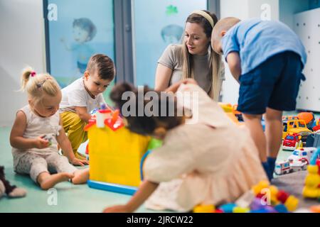 Learning through play at the nursery school. Toddlers and their teacher playing with colorful plastic playhouses, building blocks, cars and boats. Imagination, creativity, fine motor and gross motor skills development. High quality photo Stock Photo