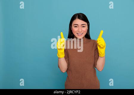 Happy optimistic young woman asian thumbs up and wearing yellow gloves for hand safety isolated on a blue background, Cleaning home concept, Excited pleasant female with positive behavior Stock Photo