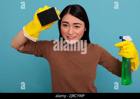 Smiling happy and joyful asian young woman holding a sponge with yellow gloves isolated on a blue background, Housewife worker, Cleaning home concept, Delighted laughing and positive state of mind Stock Photo