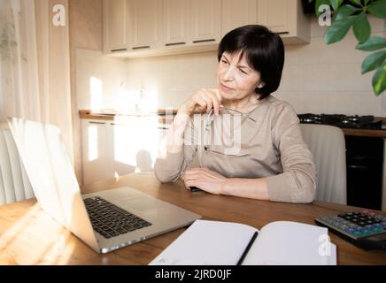 Beautiful sophisticated elderly woman holding glasses, talking on video communication on a laptop. Successful old lady smiles and works from home. Stock Photo