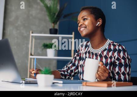 Young African American woman entrepreneur having a call, sitting at her office desk and drinking coffee. Successful entrepreneurship. Business call. Stock Photo