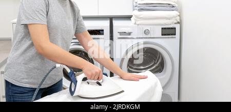 Woman ironing on board in laundry room with washing machine on background Stock Photo