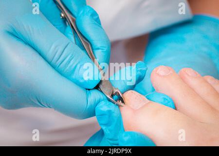 A chiropodist in blue medicine gloves gives a pedicure to the client's foot, cutting dry skin with clippers. Close up. The concept of salon foot care. Stock Photo