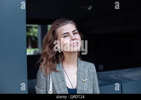 Portrait of young smiling slim green-eyed woman businesswoman standing near modern building outside, looking at camera. Stock Photo