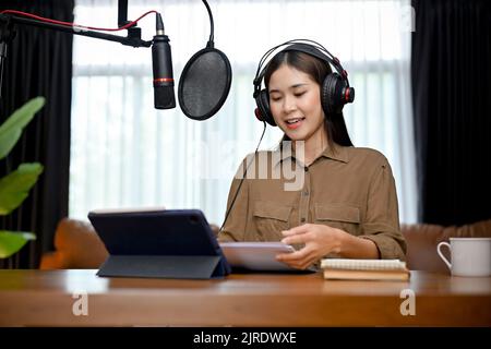 Beautiful young Asian female online radio host reading and reporting a daily morning news to her online followers, using headphones and professional m Stock Photo