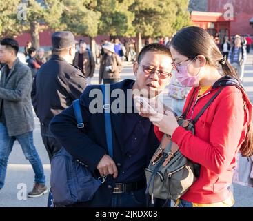 A young Chinese woman wearing a face mask and a Chinese man looking at a smartphone. standing outside the Forbidden City. Stock Photo