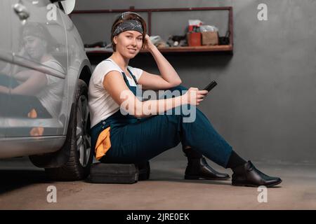 Smiling caucasian pretty woman mechanic is sitting leaning on a car in an auto repair shop and using cellphone. The concept of women's work in male pr Stock Photo