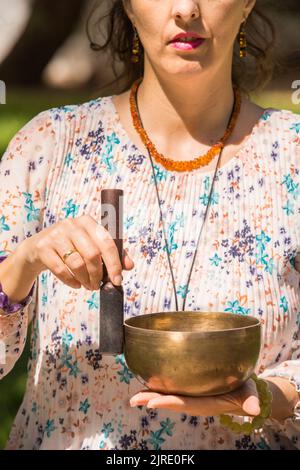 Detail of a woman's hands holding and playing a Tibetan singing bowl during a meditation and music therapy session in a park area. Stock Photo