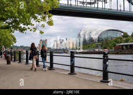 Two people walking on the Quayside, Newcastle upon Tyne, UK, under the Tyne Bridge with a view of Gateshead landmarks including The Glasshouse Stock Photo